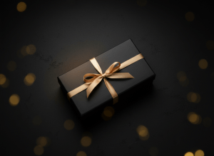 Ultra-dark luxury moody photograph, near-black charcoal background, an elegant matte black gift box with a thin satin gold ribbon softly lit from the side, subtle golden bokeh, cinematic low-key lighting, premium discreet boutique aesthetic, no text, high-end product photography